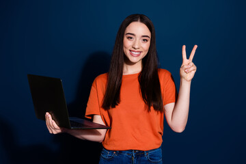 Photo of cheerful nice glad woman wear orange trendy clothes hold gadget v-sign isolated on dark...