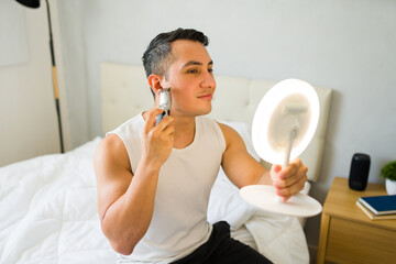 Attractive hispanic man using a ring light mirror to groom his beard on the bed