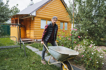 A man diligently working in his garden with a wheelbarrow situated near a charming wooden house,...