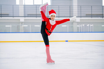 Little girl figure skater dressed as Santa Claus skates on the ice of an indoor arena.