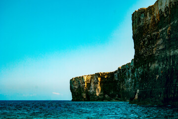 Massive coastal cliffs towering above the blue ocean under a clear sky with sunlight illuminating...