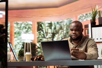 Young self employed businessman does online shopping with his card, making electronic banking payments at his home office. Freelancer does remote work and purchases things on laptop.