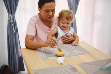 Happy toddler sitting with mother while learning to eat at the dining table with joy and family bonding