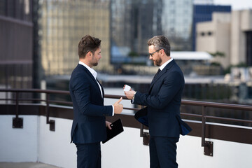 Business meeting of friends outdoors. Two men in suits are sitting on a bench near a city building with a laptop and talking. Partnership and corporate relations. Smiling partners discussing