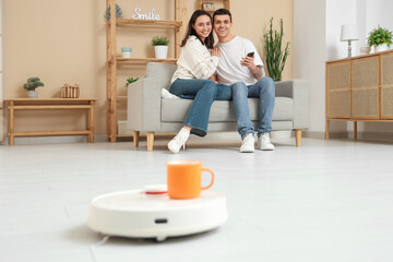 Young couple using mobile phone for remote control of modern robot vacuum cleaner with cup in living room