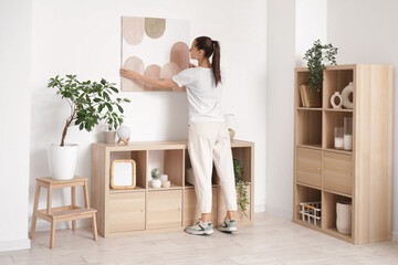 Young beautiful woman hanging picture on white wall in stylish room
