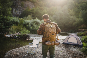 Adult man hiker with a backpack near campsite by river in forest