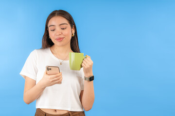 Young woman enjoying a relaxing moment, drinking coffee and checking her phone on blue background