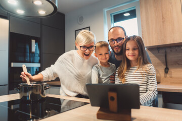 Happy family communicating with grandparents over video call while mom cooks lunch in the kitchen