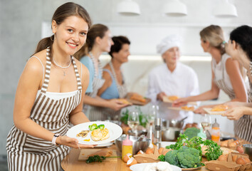 Young woman holding plate with cooked chicken breast in her hands posing surrounded by other members of cooking course.