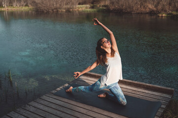 Young adult woman doing yoga by the lake in beautiful mountains. Aerial shot. Taking care of her mental health, focusing on breathing.