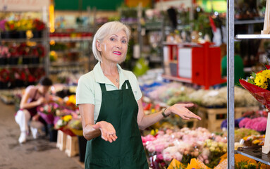 Glad aged woman seller posing against the background of colorful plants in point of sale of flowers