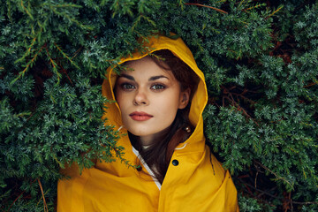 Mysterious young woman in a vibrant yellow raincoat hiding behind lush evergreen trees in a remote...