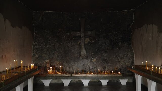 Small ancient Christian altar inside a mountain rock with burning church candles. Yellow candles burn in worship and prayer. A cross carved on the stone wall in background in greek orthodox monastery