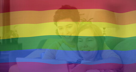 Image of pride rainbow flag over happy caucasian lesbian couple using laptop in bed