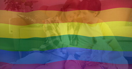 Image of pride rainbow flag over happy caucasian lesbian couple relaxing on couch with pet dog