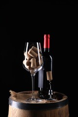 Corkscrew with metal handle, bottle of wine, glass and corks on wooden barrel against black...