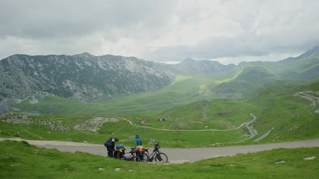 Couple preparing their bicycles for a ride downhill on windy road in Montenegro. Bike camping in the Balkan region. Two people travel together in green and picturesque scenery of mountains and valleys