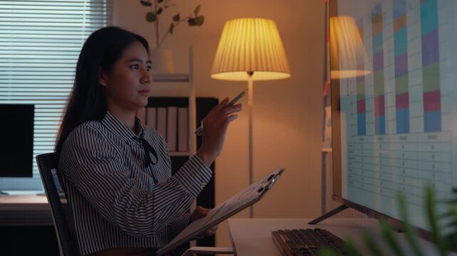 Businesswoman seated at a desk reviews a gantt chart on her computer while jotting down notes on a clipboard. Focused on project management, she works late into the evening