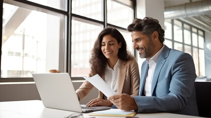 A man and woman dressed smartly, are sitting at an office desk with a laptop open as they look over...