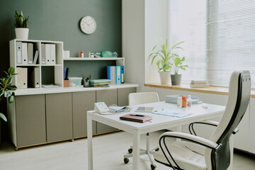 Office interior with minimalist decor featuring organized shelves, a desk with office supplies, potted plants, and large windows allowing natural light into workspace