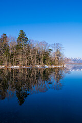 晴れた日の冬の湖畔。森の葉の無い木々が水面に反射して美しい冬景色を描いている。日本の北海道の屈斜路湖。
