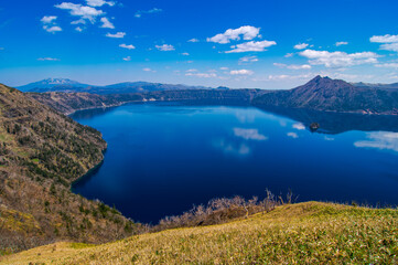 雲の浮かぶ青空を反射する山間の湖。日本の北海道の摩周湖は美しい自然の景色が観光お客に人気の名所。雄大で迫力のあるカルデラ湖の風景が魅力。