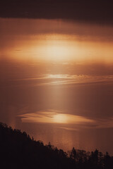 広大な湖面に太陽の光が反射し、水面全体が黄金色に輝いている。遠方にはシルエットになった山々が連なり、雄大な自然の風景が広がる。金色の光が湖を照らし、神秘的な雰囲気を醸し出す。