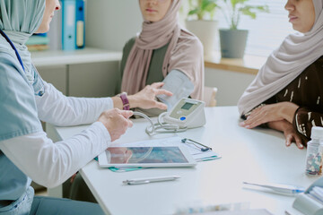 Healthcare professional speaking with veiled patient about medical results while using stethoscope and blood pressure monitor during consultation in office setting