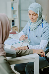 Obraz premium Medical professional wearing hijab attending to patient in modern clinic setting, performing checkup. Stethoscope and medical documents present, conveying professionalism and care