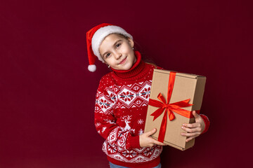 Cheerful child in a festive red sweater and Santa hat holding a gift box against a deep red background during the holiday season