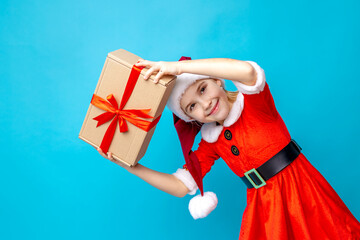 Obraz premium Cheerful girl in a red Christmas dress and Santa hat holds a gift box while smiling against a bright blue background during holiday celebrations