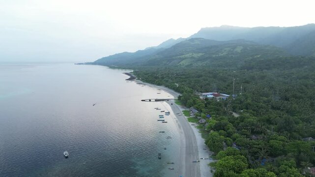 Dili coastline with lush greenery, beach, and distant mountains at sunrise, aerial view