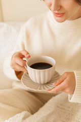 Close-up of a young woman in a cozy, light-colored outfit drinking coffee in bed at home.