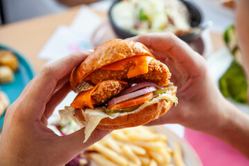 Hands holding fried chicken burger in brioche potato bun in queso sauce.