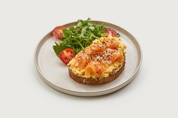 Breakfast plate featuring a slice of whole-grain toast topped with scrambled eggs, smoked salmon, and sesame seeds. A fresh side salad of cherry tomatoes and greens adds a vibrant touch.
