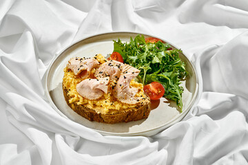 Breakfast plate featuring a slice of whole-grain toast topped with scrambled eggs, smoked turkey, and sesame seeds. A fresh side salad of cherry tomatoes and greens adds a vibrant touch.