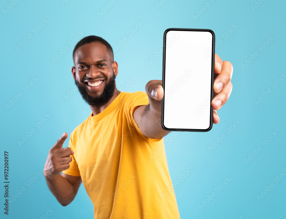 Aufkleber Excited black guy in casual wear pointing at cellphone with empty screen on blue studio background, space for website or mobile app design. Cellphone display mockup. Selective focus