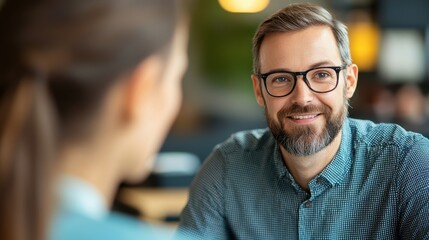 Obraz premium A smiling man with glasses engages in conversation at a modern cafe, showcasing warmth and professionalism in a relaxed setting.