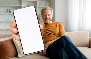 An older man relaxes on a couch in a well-lit room, smiling at the camera while holding up a blank smartphone. The setting suggests comfort and modern technology.