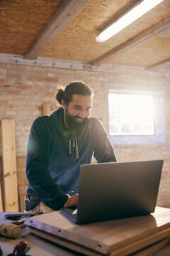 Smiling woodworker looks down at his laptop in his workshop. He emails with customers
