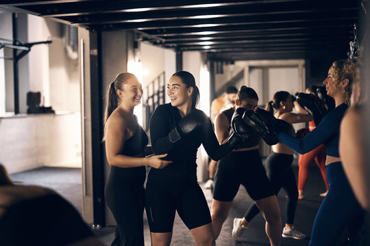 Young female coach helping a woman wearing boxing gloves laughing together while doing a punching workout during a boxing class in a gym
