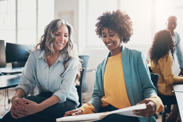 Diverse businesswomen smiling while going over paperwork in an office. Behind them, stand two colleagues talking at a computer