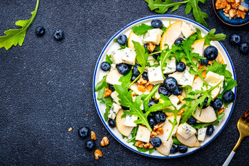 Tasty salad with pears, blueberries, roquefort cheese, arugula and nuts. Black background, top view