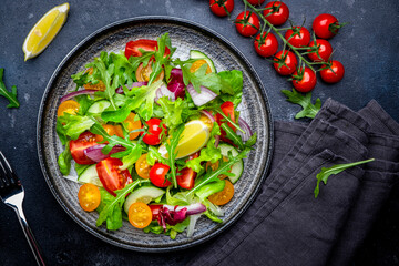 Vegan vegetable salad with tomatoes, cucumber, red onion, lettuce and arugula. Black background, top view