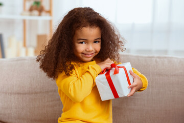 Kid's Birthday. Cute African Little Girl Unpacking Wrapped Gift Sitting With Mother On Sofa At Home.