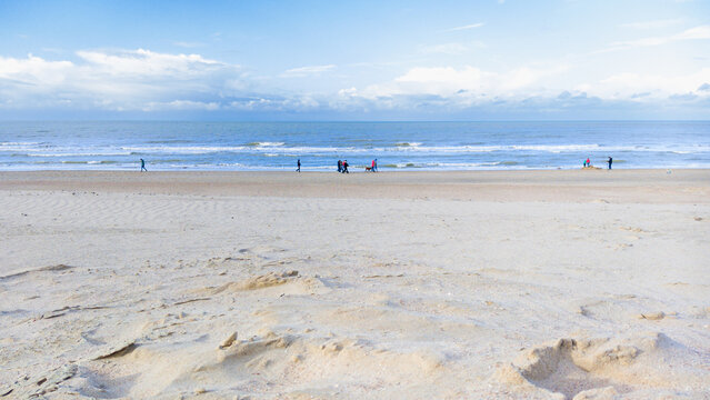 A serene coastal scene showing a wide sandy beach during low tide with a few people strolling under a cloudy sky in autumn.