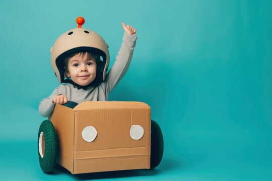 A little boy in a cardboard car wearing a helmet. He's smiling and giving a thumbs up with a joyful expression. He's enjoying his imaginative play.