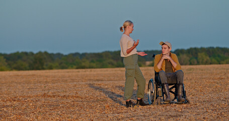 A woman chats with a young man in a wheelchair in a wide open field at sunset.