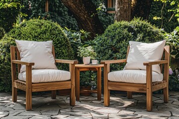 Obraz premium Two wooden garden armchairs with white cushions and a small round table between them on a stone floor, set against green bushes. Outdoor coffee set nearby creates a cozy, elegant, and relaxed atmosphe